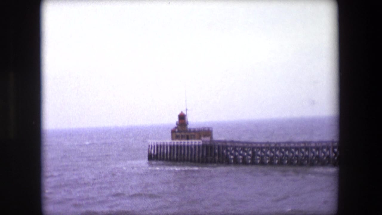 1967 Calais 8mm frame showing a small lighthouse on a long pier against choppy water and an overcast horizon.