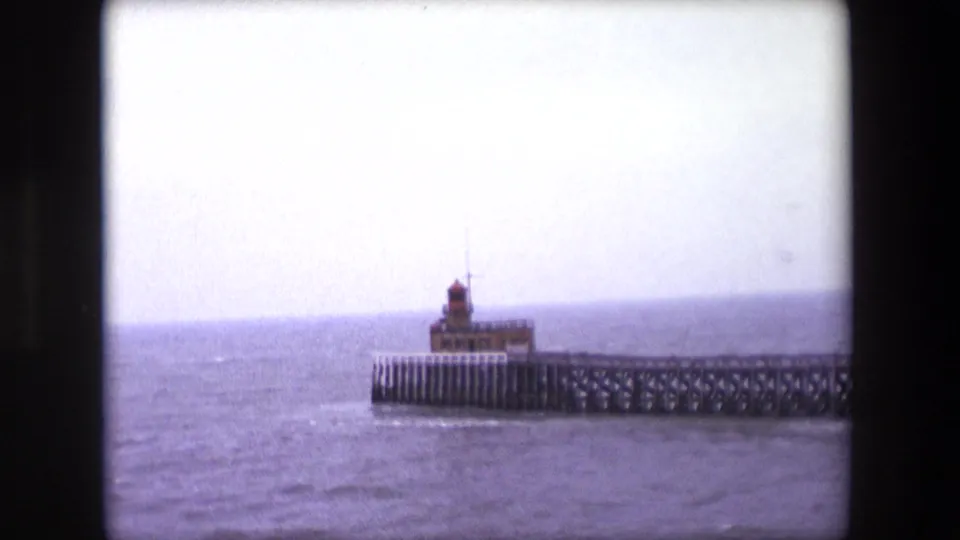 1967 Calais 8mm frame showing a small lighthouse on a long pier against choppy water and an overcast horizon.