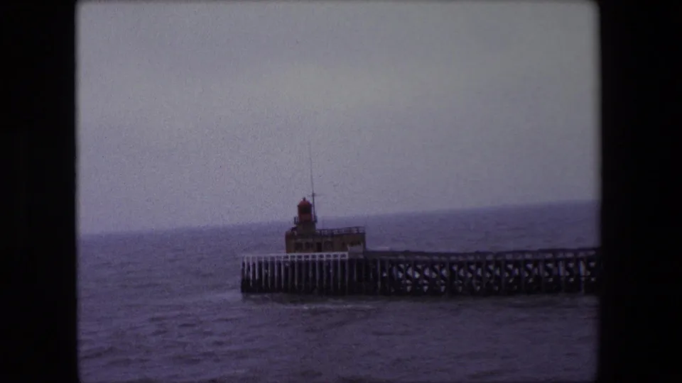 1967 Calais 8mm frame with the lighthouse centered near the end of the pier, surrounded by muted sea and cloudy sky.