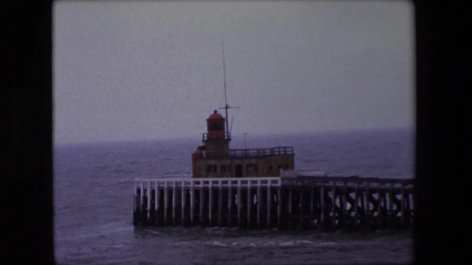 1967 Calais 8mm frame with a closer view of the pier-end lighthouse, railings, pilings, and choppy coastal water.