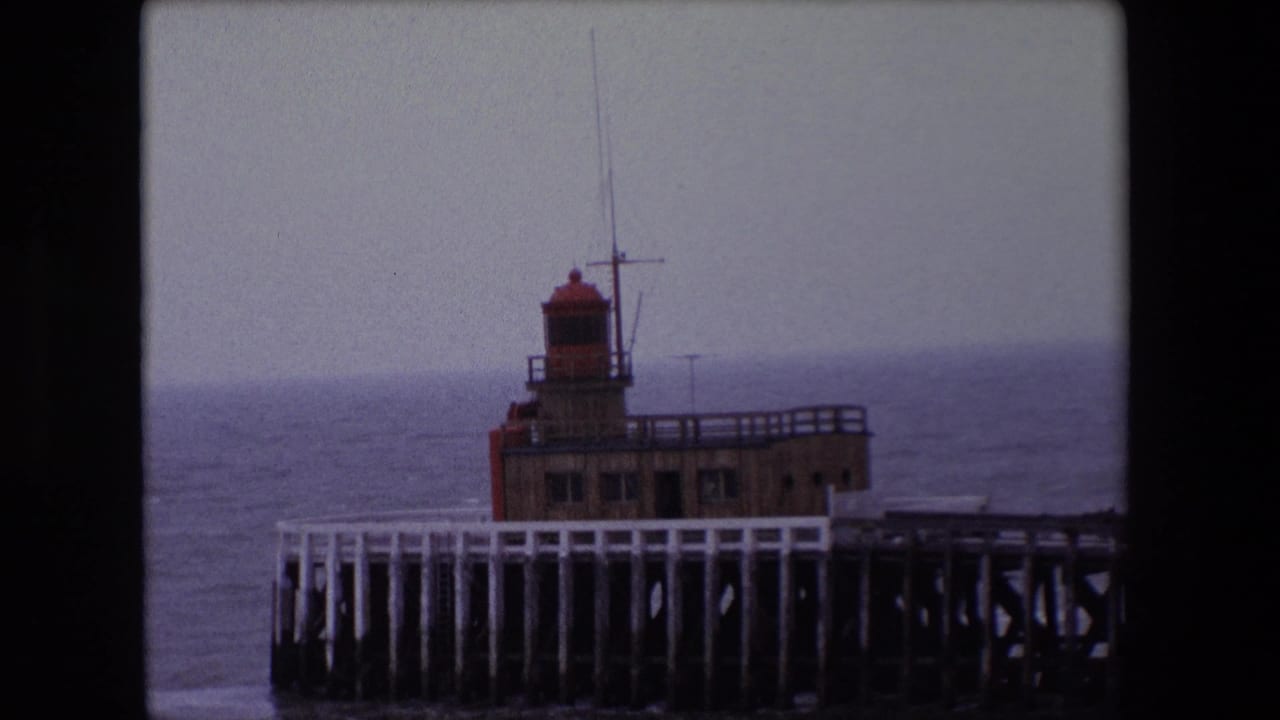 1967 Calais home-movie frame showing a red-topped lighthouse structure on a wooden pier against an overcast sea horizon.