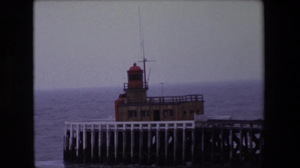 1967 Calais home-movie frame showing a red-topped lighthouse structure on a wooden pier against an overcast sea horizon.
