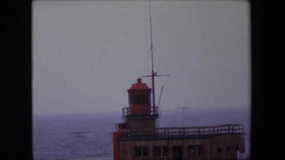 Closer 1967 Calais view of the red lantern room, antenna mast, and pier railing with gray-blue water in the background.