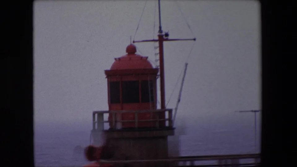 Tight 1967 Calais home-movie view of the red lighthouse lantern room, mast, and surrounding railing set against a hazy sea and sky.