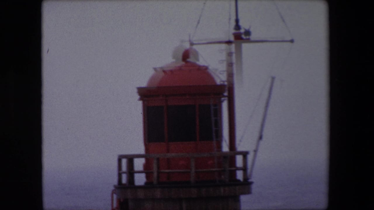 1967 Calais home-movie frame showing a red lighthouse lantern room, wooden railing, and mast structure against a pale foggy sky.