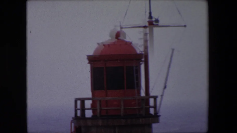 1967 Calais home-movie frame showing a red lighthouse lantern room, wooden railing, and mast structure against a pale foggy sky.