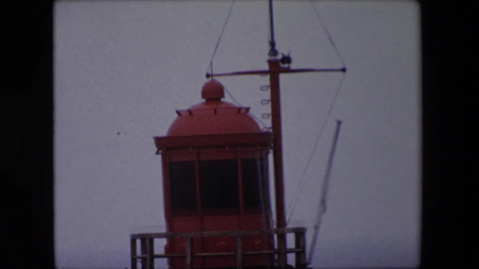 1967 Calais footage frame with the upper red lighthouse structure partly cropped, a mast, rigging lines, and muted foggy sky.