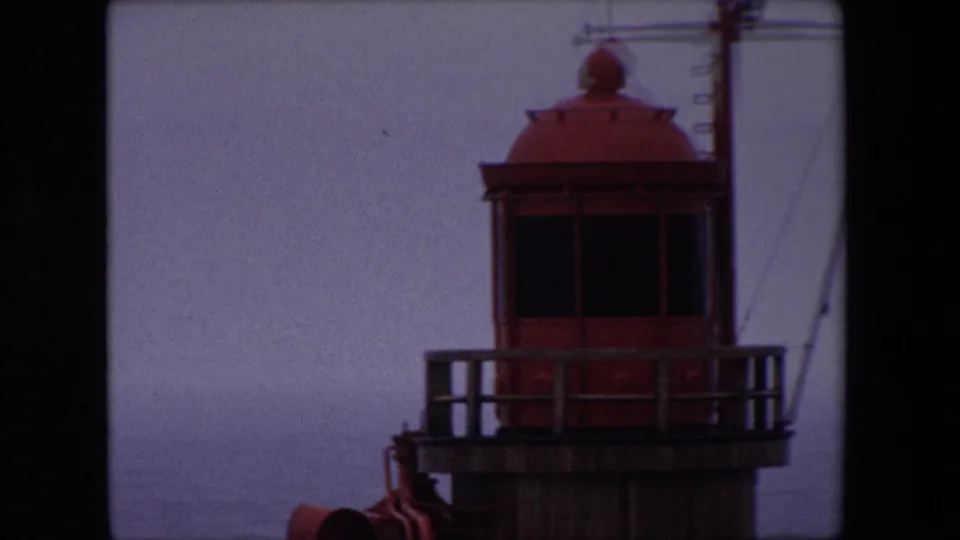 Closer 1967 Calais frame of the red lighthouse lantern room, dark windows, platform railing, and mast details in soft 8mm focus.