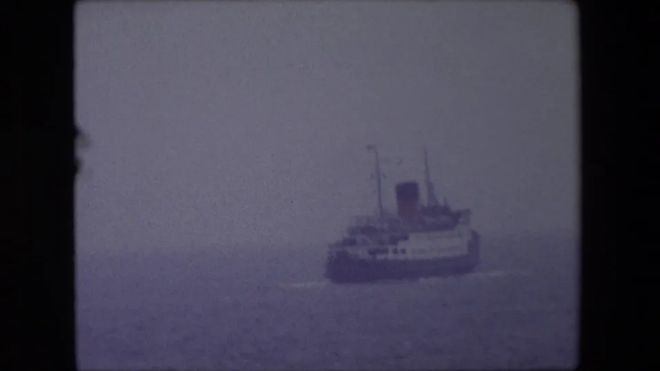 The vessel continues across calm water near Calais, its hull, decks, funnels, and wake softened by fog and 8mm film grain.