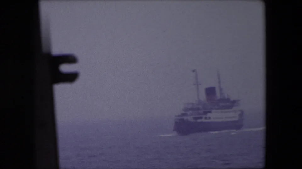 The ship appears farther right in the 8mm frame, leaving a faint wake across calm water under the hazy Calais sky.