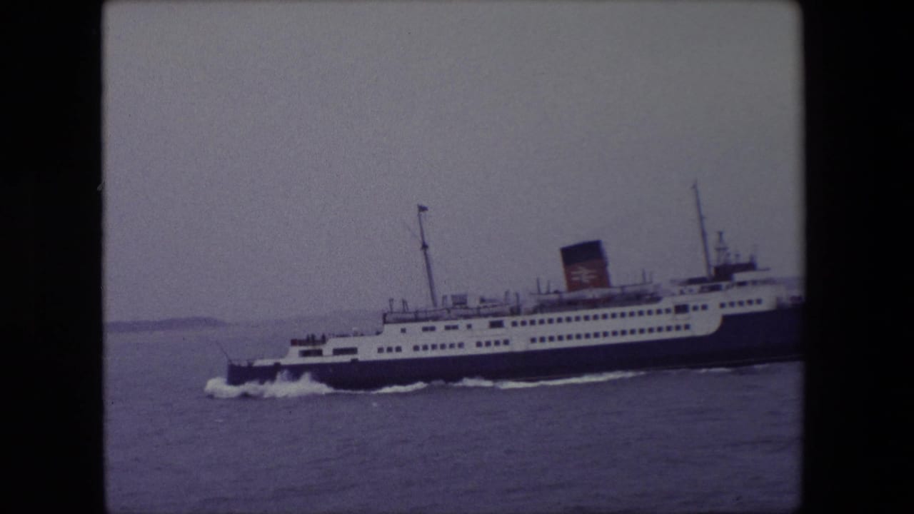 1967 Calais home-movie frame showing a large white and dark blue passenger ferry crossing grey water under an overcast sky.