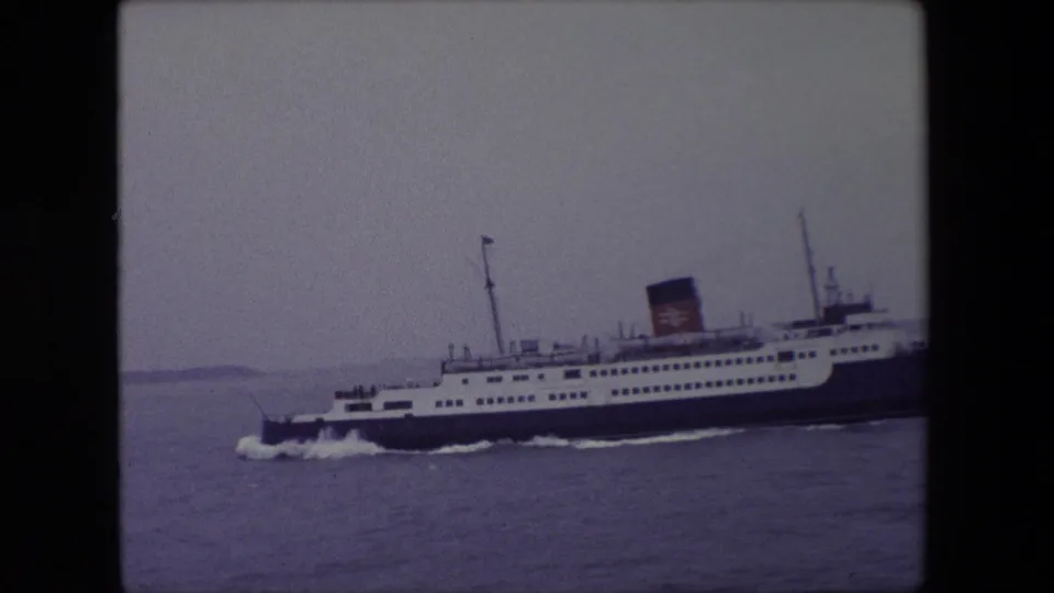 1967 Calais home-movie frame showing a large white and dark blue passenger ferry crossing grey water under an overcast sky.