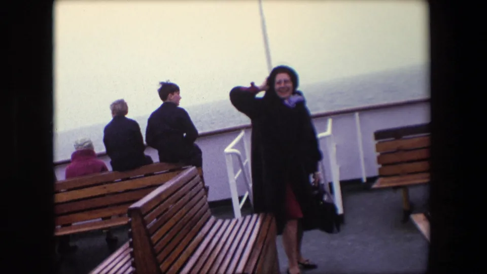 1967 Calais ferry deck scene with passengers near wooden benches, including a woman in a dark coat and hat facing the camera, with hazy sea beyond the railing.