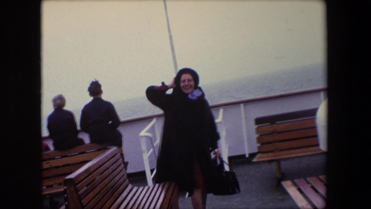 In Calais, France, 1967, a woman in a dark coat and hat smiles on a windy ship deck with seated passengers, wooden benches, white railings, and sea behind her.