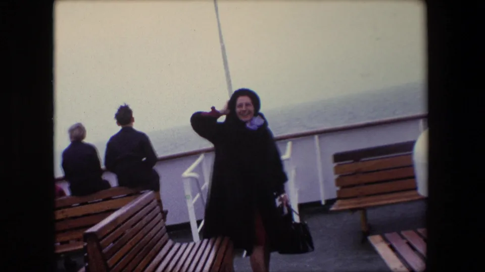 In Calais, France, 1967, a woman in a dark coat and hat smiles on a windy ship deck with seated passengers, wooden benches, white railings, and sea behind her.