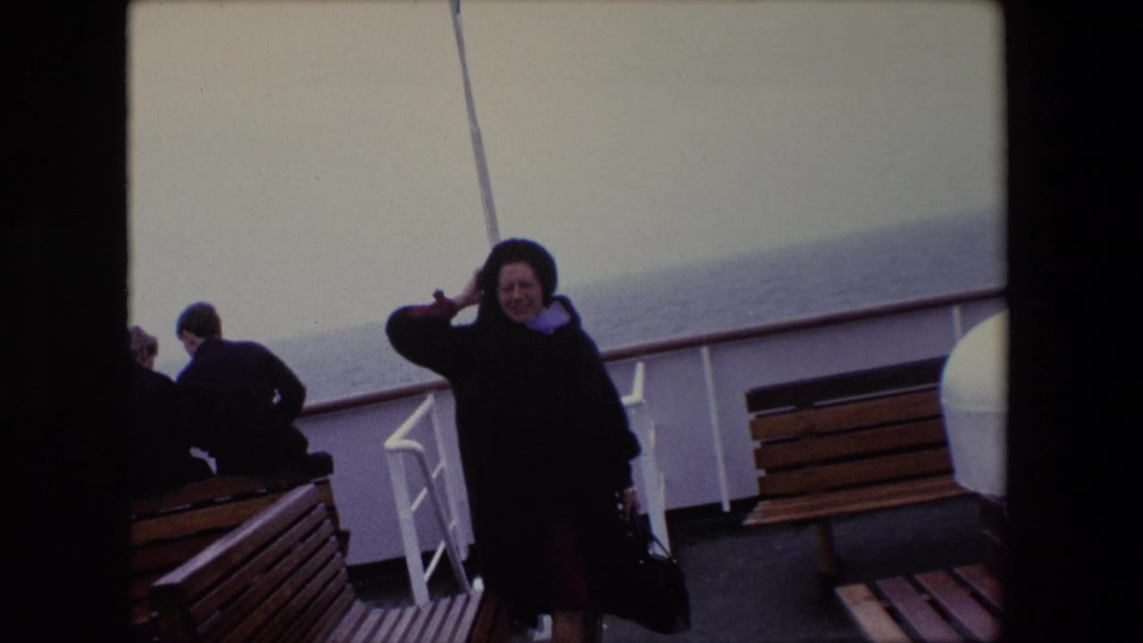 Woman holds her hair or hat while standing on a ship deck with benches and sea in the background.