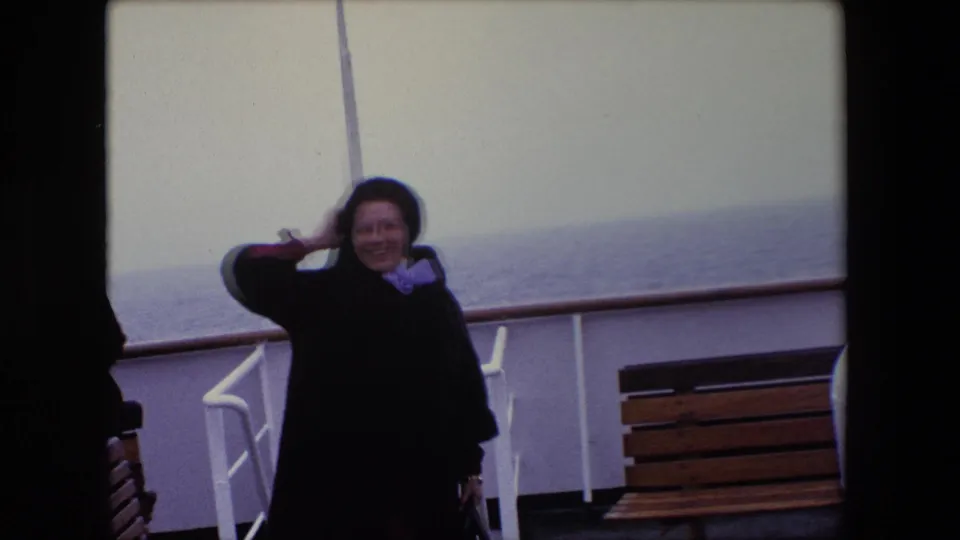 A woman in a dark coat smiles toward the camera on a windy ship deck, with a wooden bench, white railings, and the sea visible in the Calais 1967 home-movie frame.