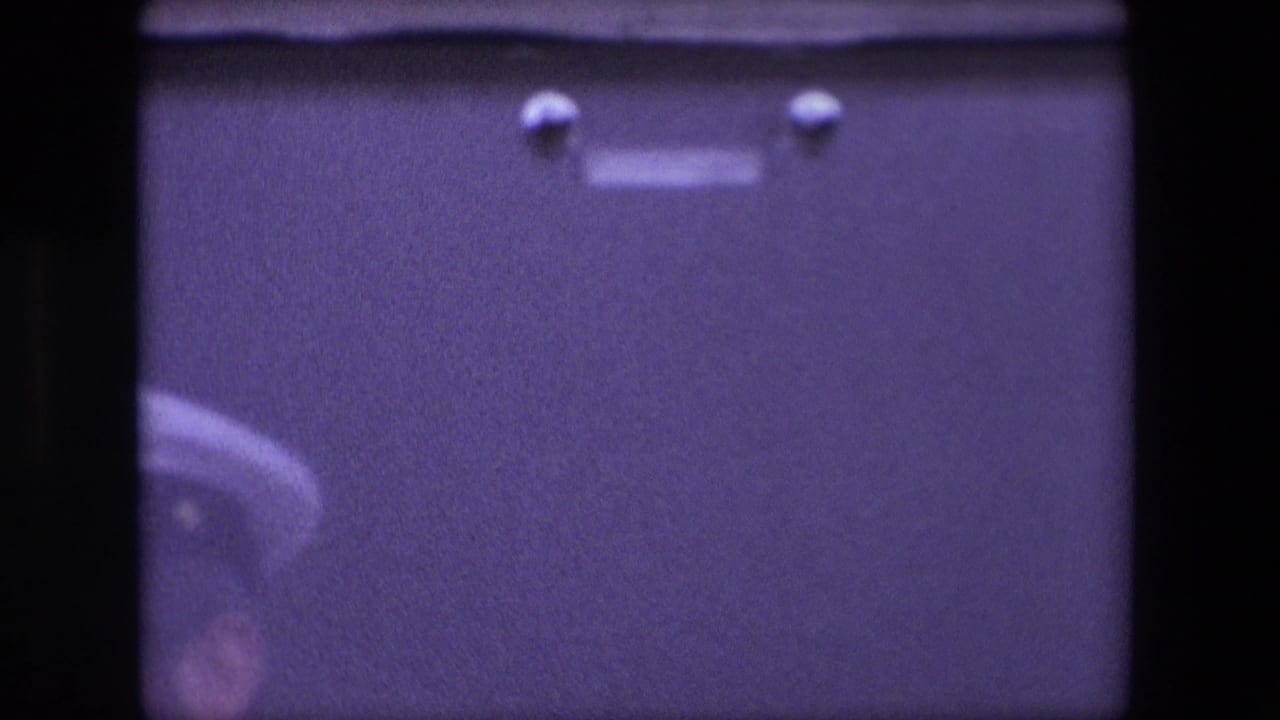 Soft, purple-tinted 8mm frame from the Calais 1967 clip showing an indistinct white-capped uniformed figure near a dark vertical film edge.