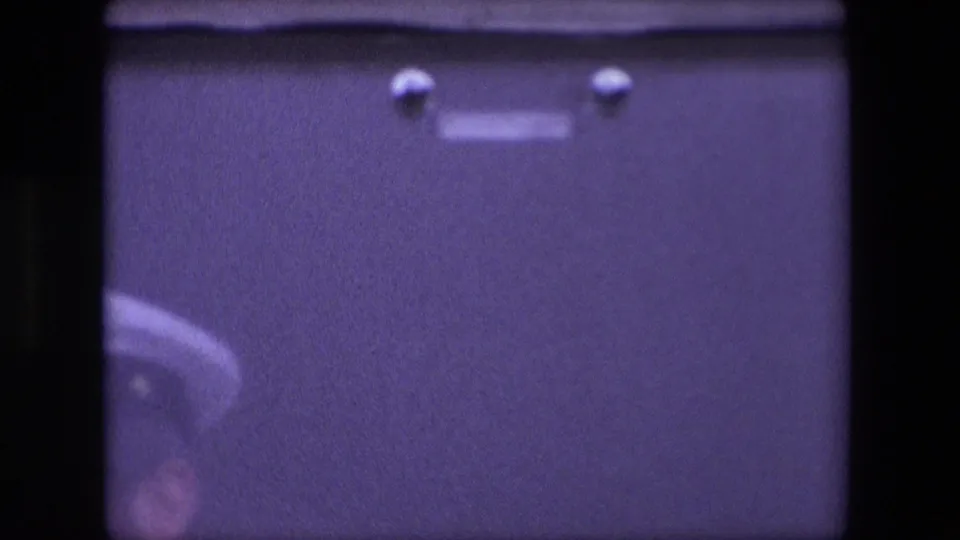 Soft, purple-tinted 8mm frame from the Calais 1967 clip showing an indistinct white-capped uniformed figure near a dark vertical film edge.
