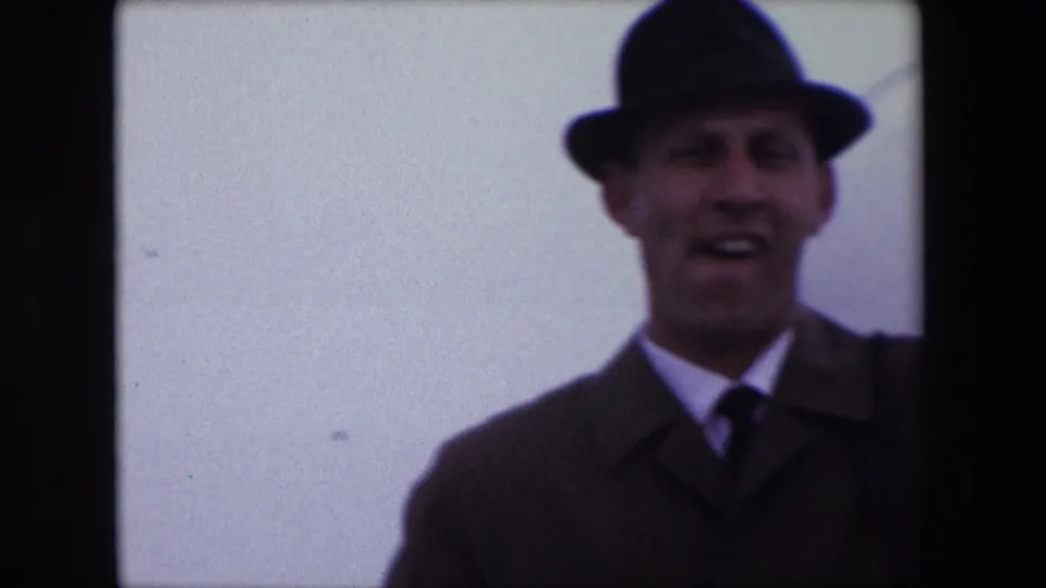 In the Calais 1967 home-movie frame, an adult man in a dark fedora, coat, shirt, and tie appears to speak toward the camera against an overexposed background.