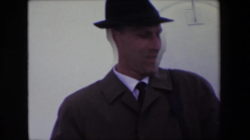 Adult man in a dark fedora and coat turns to the side in the Calais 1967 8mm frame, with a pale overexposed background and faint curved marking behind him.