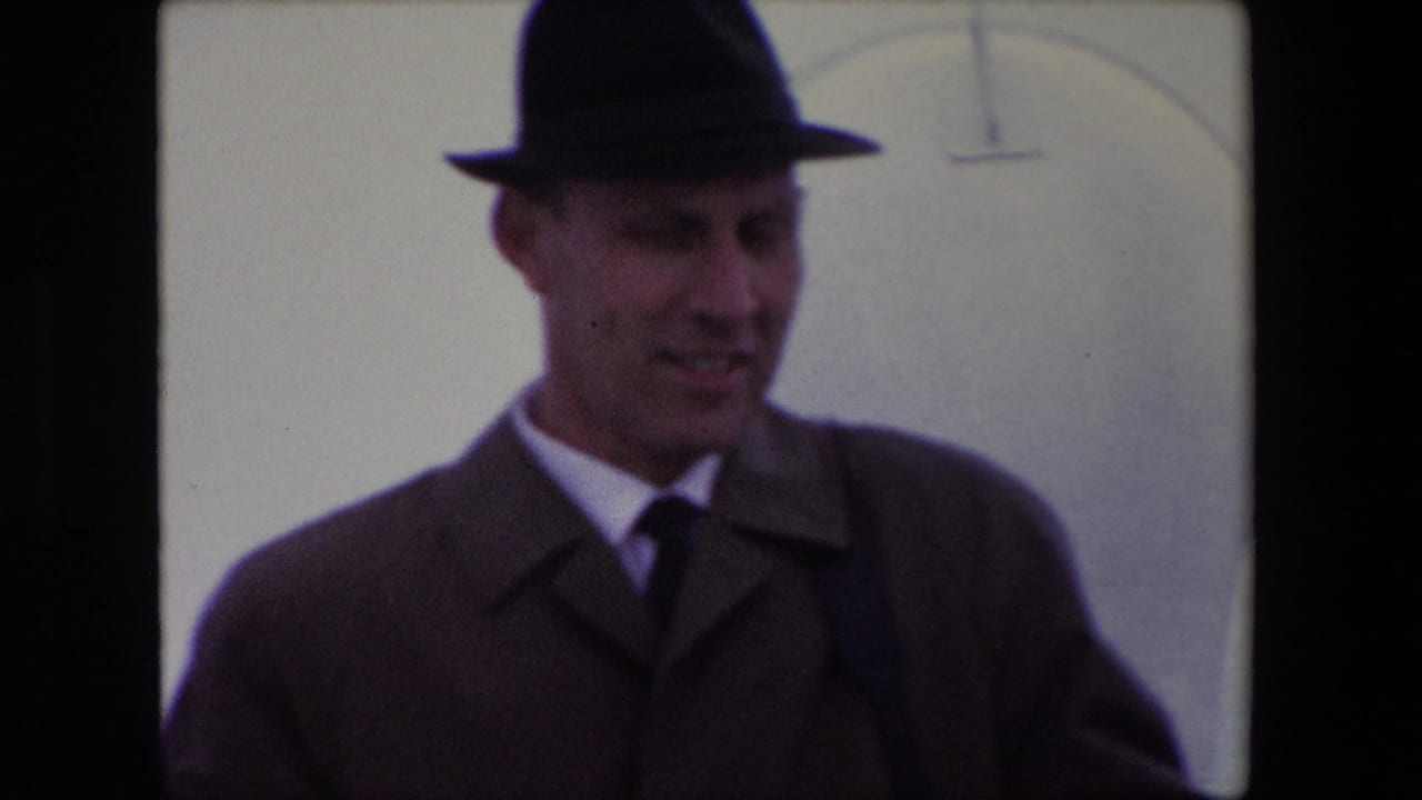 In supplied 1967 Calais home-movie footage, an adult man in a dark fedora, tie, and overcoat looks downward beside a pale wall-like background.