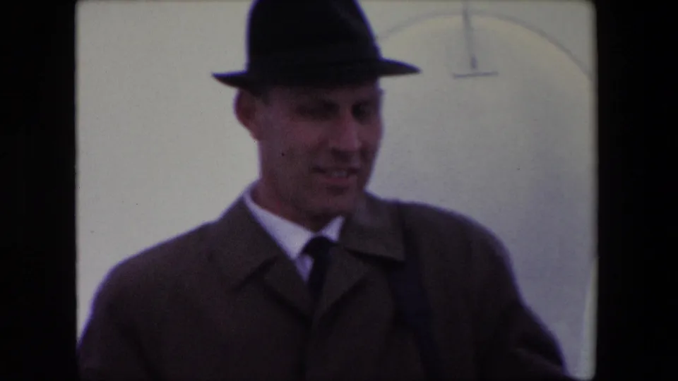 In supplied 1967 Calais home-movie footage, an adult man in a dark fedora, tie, and overcoat looks downward beside a pale wall-like background.
