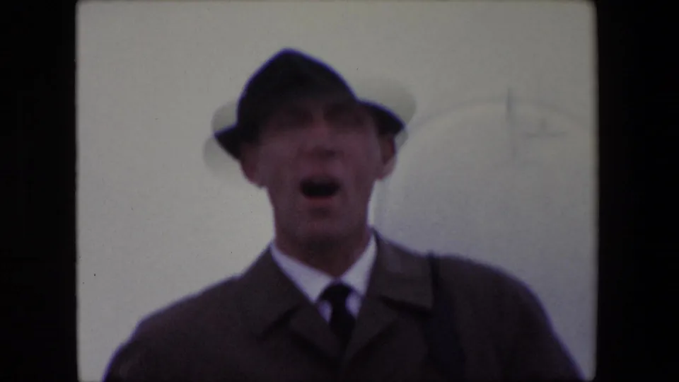In supplied 1967 Calais footage, the man faces the camera in a fedora and overcoat with his mouth open, as if speaking or laughing.