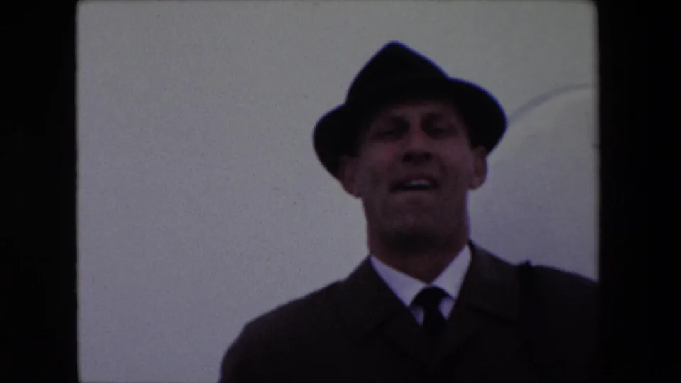 The 1967 Calais home-movie frame shows a closer view of the adult man in fedora, overcoat, shirt, and tie smiling toward the camera.