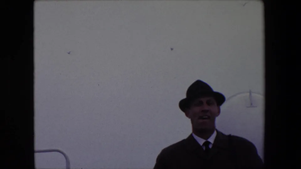 A wider supplied 1967 Calais frame shows the man smaller in view, wearing a fedora and dark coat against a pale background with a partial curved fixture or marking.
