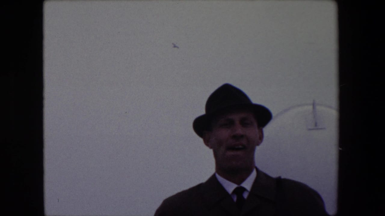 A man in a dark suit, tie, and fedora appears to speak toward the camera in 1967 Calais home-movie footage, with a pale overcast sky and faint curved structure behind him.