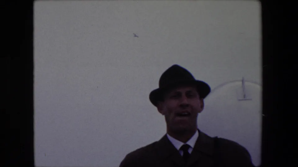 A man in a dark suit, tie, and fedora appears to speak toward the camera in 1967 Calais home-movie footage, with a pale overcast sky and faint curved structure behind him.