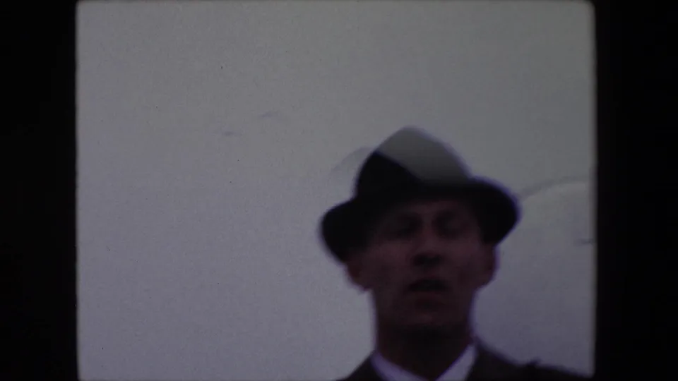 The same fedora-wearing man is seen in softer focus near the right edge of the frame, with his mouth open as if speaking and a mostly blank overcast sky behind him.