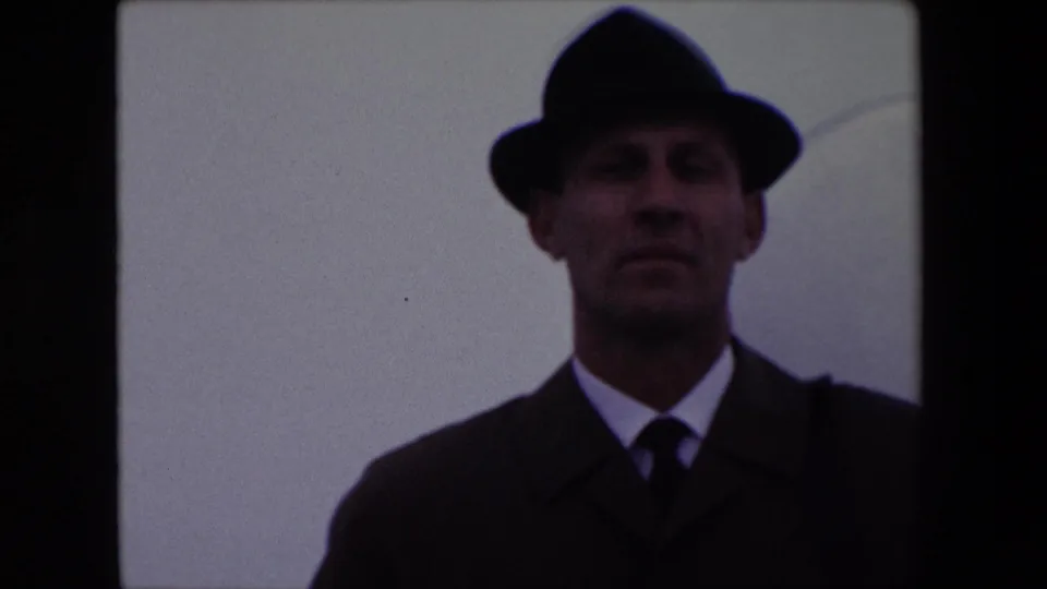 A closer, soft 8mm frame shows the man facing the camera in a dark fedora, coat, white shirt, and tie, with minimal background detail beyond the pale sky.