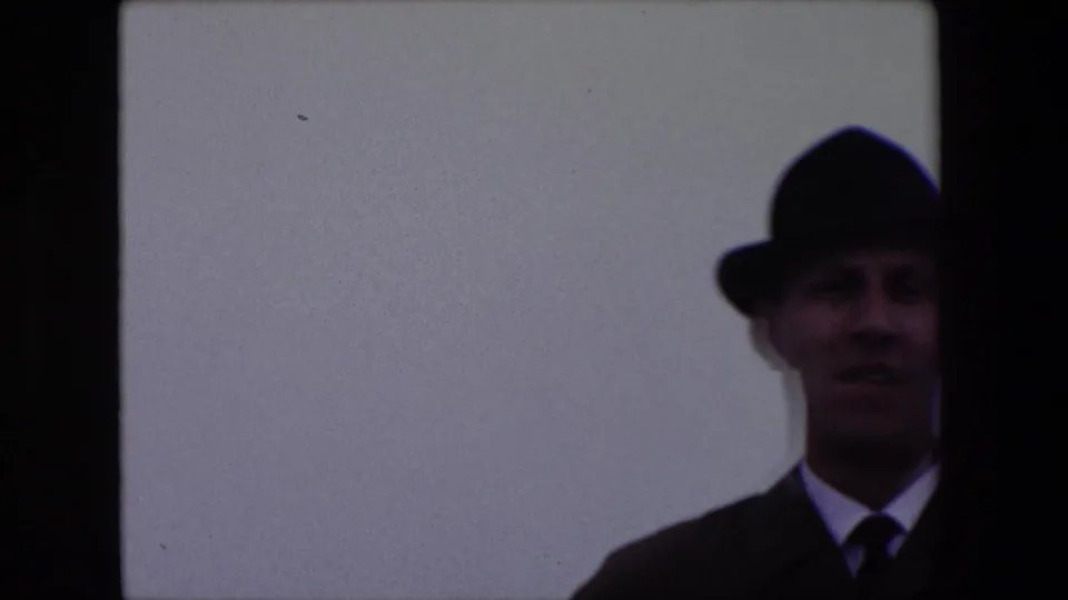 The man appears partly cropped on the right side of the frame in his dark fedora and suit, still outdoors against an empty pale sky in the short 1967 Calais clip.
