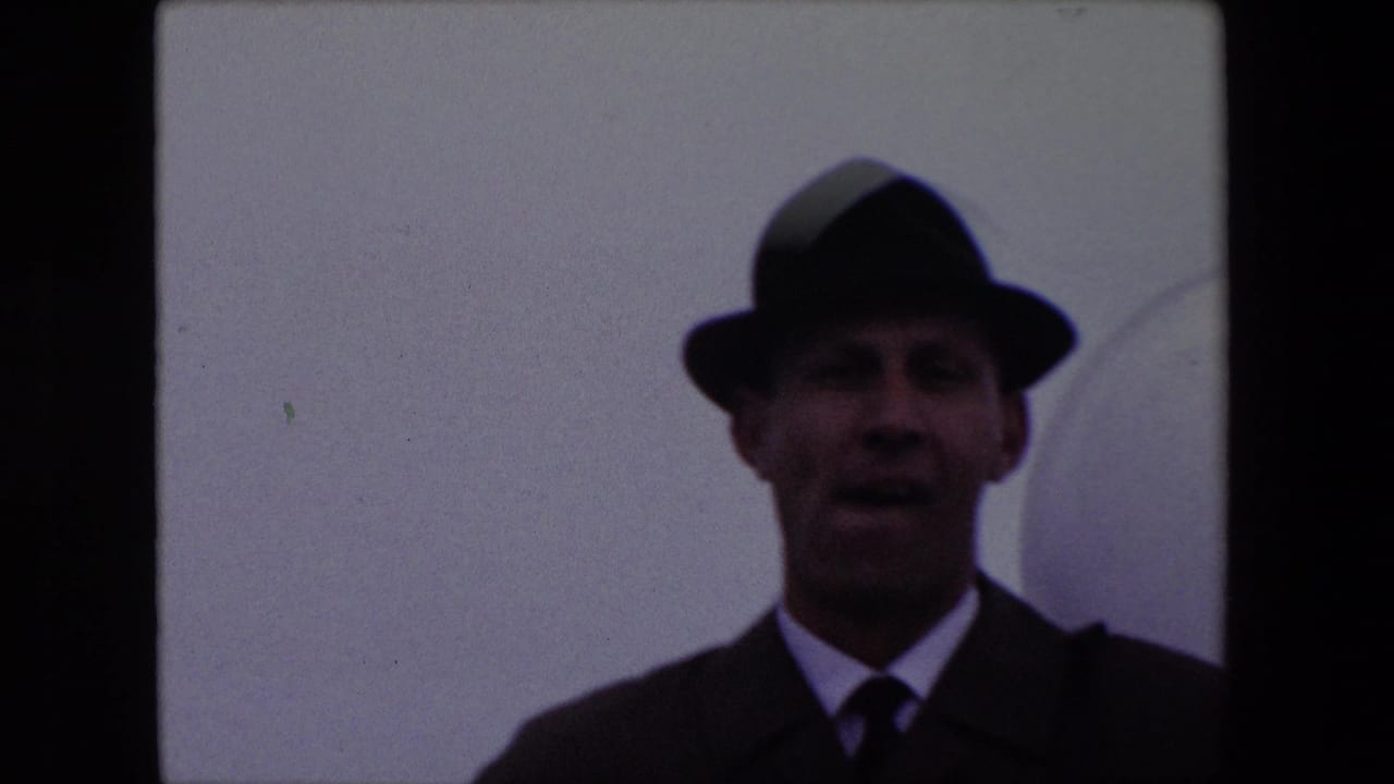 Adult man in a dark fedora and suit facing the camera against a pale wall.