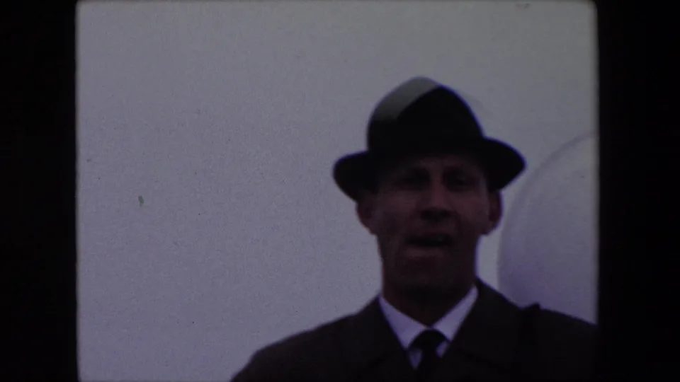 In this 1967 Calais home-movie frame, an adult white man in a dark fedora, suit jacket, white shirt, and tie faces the camera in soft focus against a plain light wall.