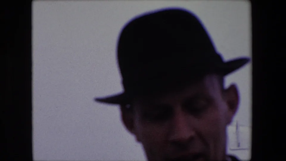 In this Calais, France 1967 home-movie frame, a shadowed adult man in a dark fedora leans close to the camera, with the film gate and window division visible around him.