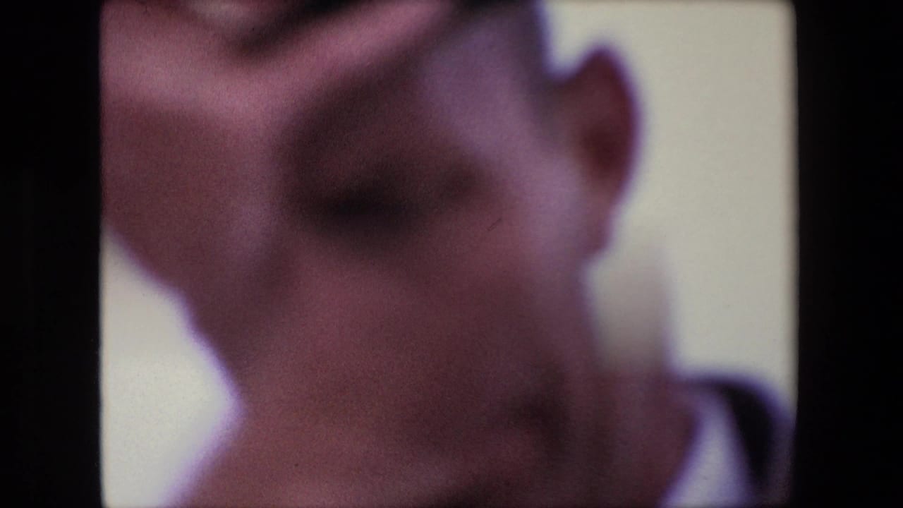 A very soft, close home-movie view shows an adult face near the camera, with facial detail obscured by motion blur, exposure, and 8mm grain.