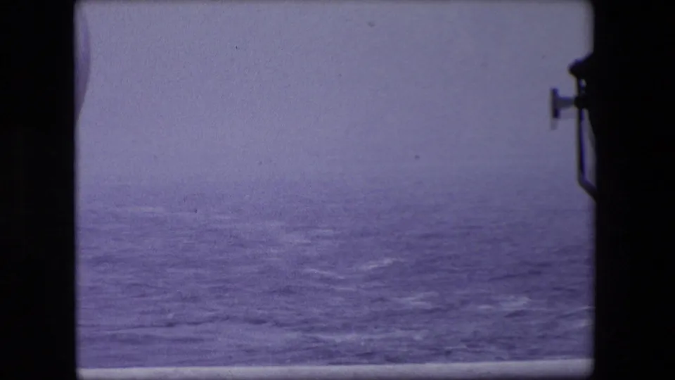 A hazy view of open water appears through a ship window, with the dark interior frame and washed 8mm color reinforcing the passenger viewpoint.