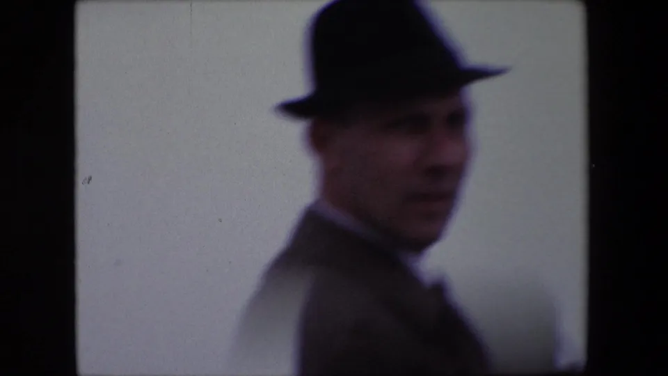 In metadata-supported 1967 Calais footage, a man in a dark fedora and suit turns near the camera in soft focus against a pale sky or sea backdrop.