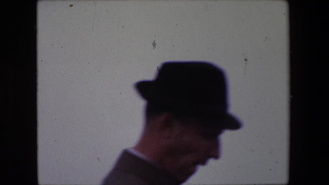 A suited man in a dark hat appears in side profile in the 1967 Calais home-movie footage, with his face partly visible against a washed pale backdrop.