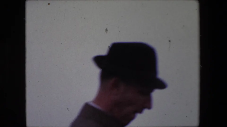 A suited man in a dark hat appears in side profile in the 1967 Calais home-movie footage, with his face partly visible against a washed pale backdrop.
