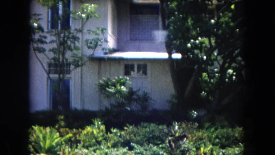 1967 Hawaii Super 8 frame showing a building corner with a white overhang, windows, palms, and thick tropical landscaping in the foreground.