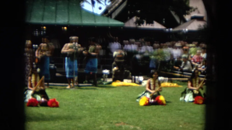 1967 Honolulu Super 8 frame showing women performing hula on a lawn while a band and a crowd watch from behind.