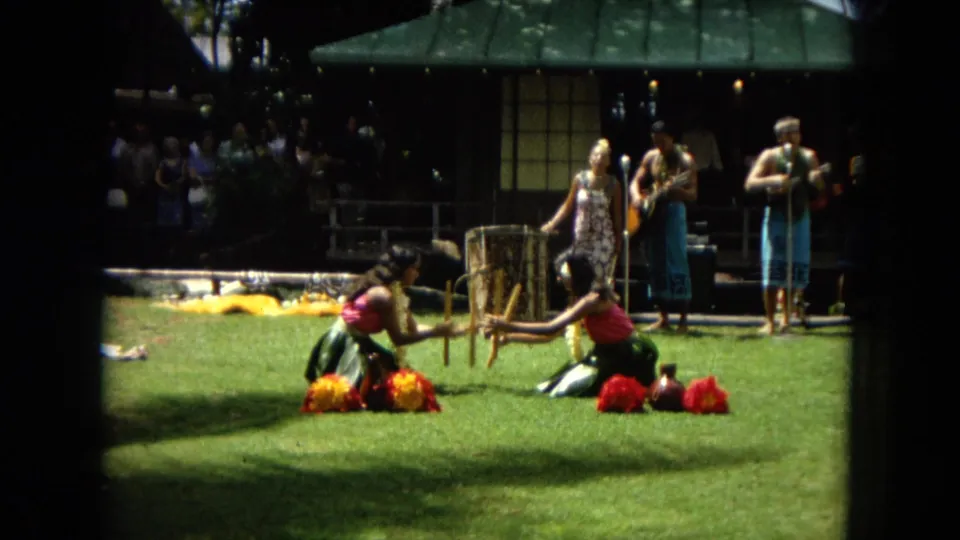 1967 Honolulu Super 8 frame showing two hula dancers seated on the grass using wooden implements while a small band performs behind them.