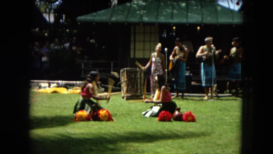 1967 Honolulu home-movie frame of hula dancers seated on grass, with musicians, a shaded structure, and spectators visible in the background.