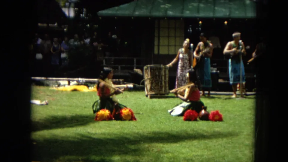 1967 Hawaii Super 8 frame showing women performing hula on a lawn while musicians play behind them and a crowd watches from the edges.