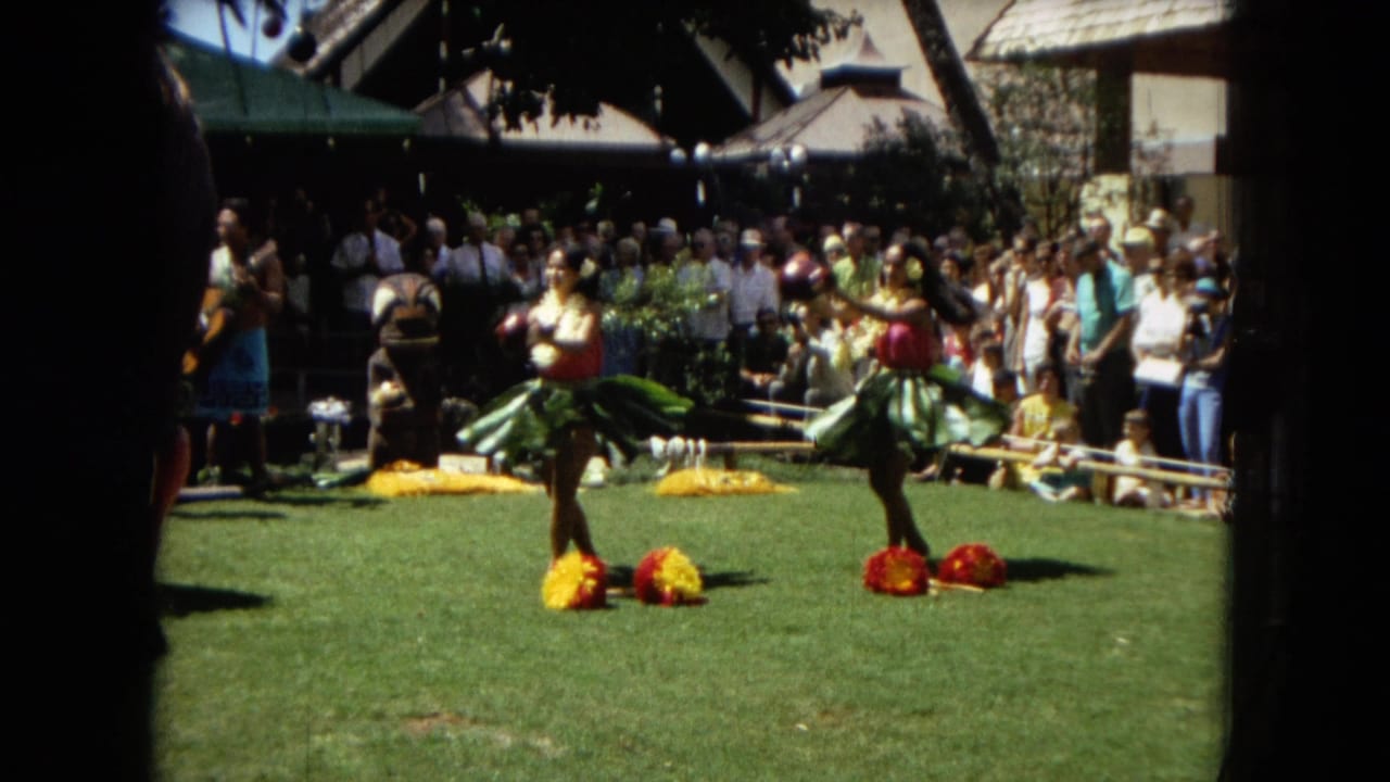 Two hula dancers stand on grass with bright floral props and a crowd in the background.