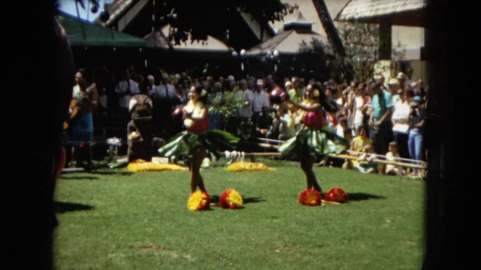 1967 Hawaii Super 8 frame with two hula dancers in green skirts performing on grass before spectators and musicians at an outdoor entertainment area.